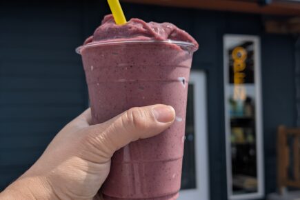 Hand holding a purple berry smoothie with a yellow straw in front of the Vim & Vigor storefront in Manette, with the café’s sign visible in the background on a sunny day.
