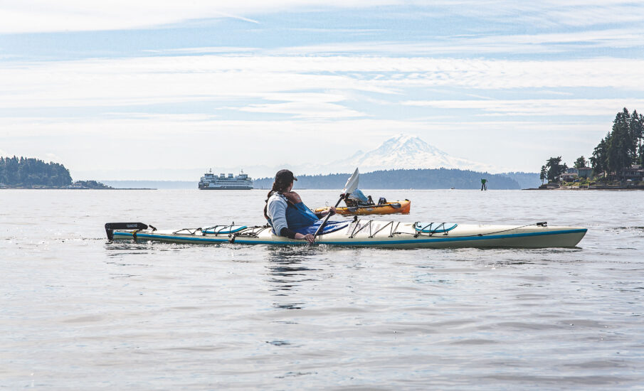 wo kayakers paddle on calm Puget Sound waters, with a Washington State ferry and Mount Rainier/Tahoma visible in the distance under a bright, cloudy sky.