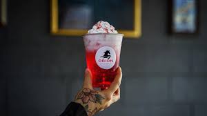 Hand holding a clear cup of bright red iced drink topped with whipped cream and an Orion Coffeehouse logo, set against a dark interior wall inside the café.