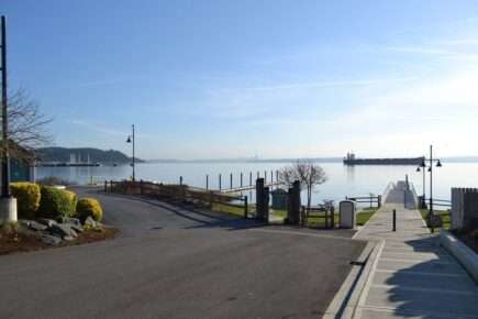 An accessible waterfront dock and boat launch at the Port of Manchester overlook calm Puget Sound, with benches, signage, and a cargo ship passing in the distance under clear skies.