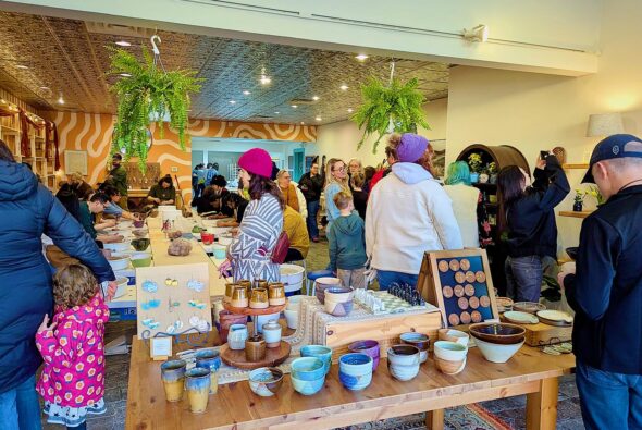 A lively pottery studio filled with people painting ceramics at long tables, finished bowls and mugs displayed in the foreground, hanging plants overhead, and families browsing and creating together in a bright, welcoming space.