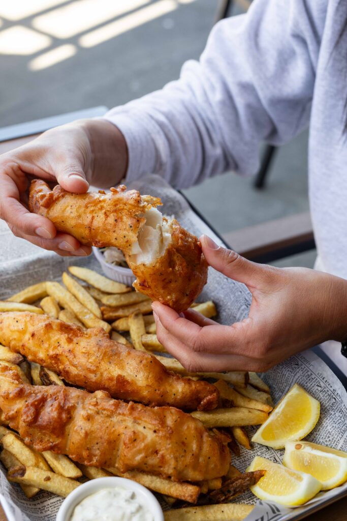 Hands breaking open a piece of golden fried fish over a basket of fish and chips, showing flaky white fish inside, with fries, lemon wedges, and tartar sauce on the side.