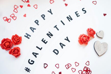 Flat lay on a white background with letter tiles spelling “# GALENTINES DAY,” surrounded by small red heart confetti, red rose decorations, and two wooden heart shapes.