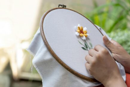 Person hand-embroidering a small yellow and white flower on fabric in a wooden hoop, with soft natural light and greenery in the background.