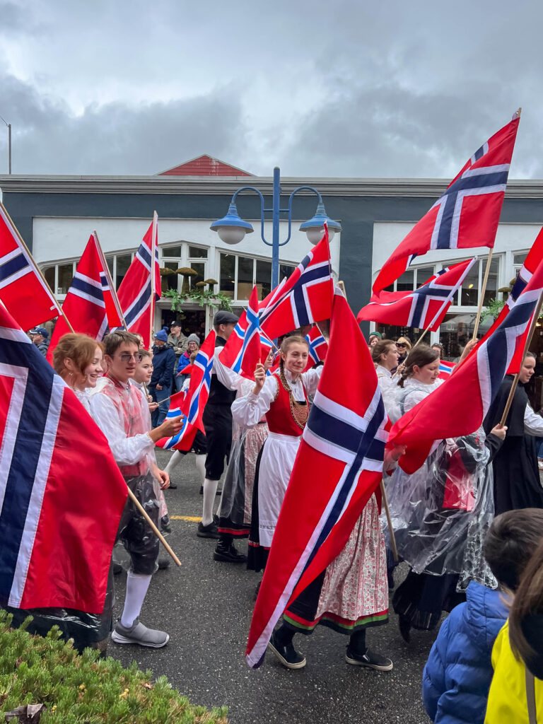 Participants in traditional Norwegian attire march in a parade in Poulsbo, Washington, waving Norwegian flags during Viking Fest, with spectators lining the street on an overcast day.