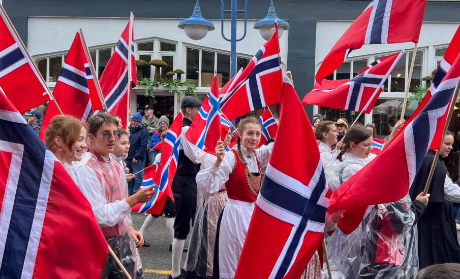 Participants in traditional Norwegian attire march in a parade in Poulsbo, Washington, waving Norwegian flags during Viking Fest, with spectators lining the street on an overcast day.