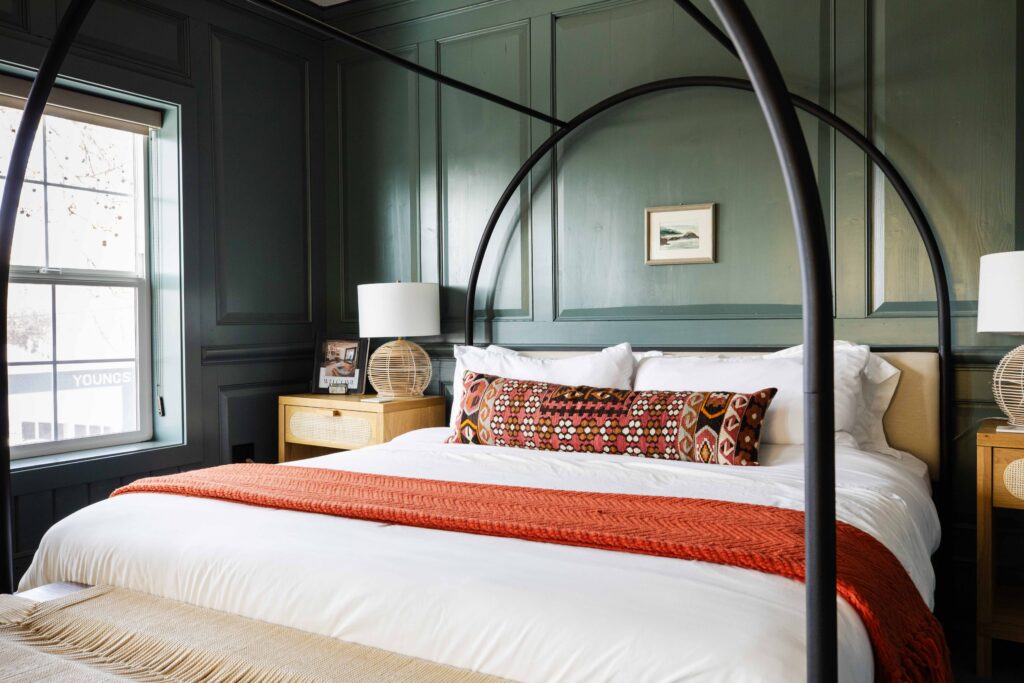 Stylish boutique hotel bedroom in Kingston, Washington featuring a canopy bed with white linens, colorful accent pillows, wood nightstands, and natural light from a window.
