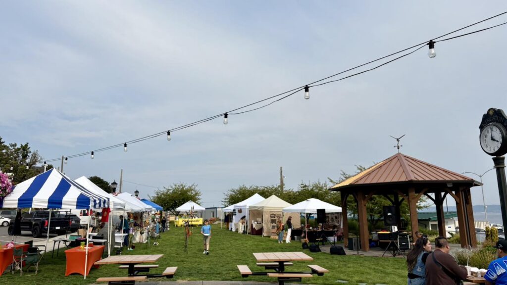 Outdoor market in Kingston, Washington featuring rows of white vendor tents, string lights overhead, and a gazebo stage near the waterfront, with visitors browsing local goods on a grassy lawn by Puget Sound.