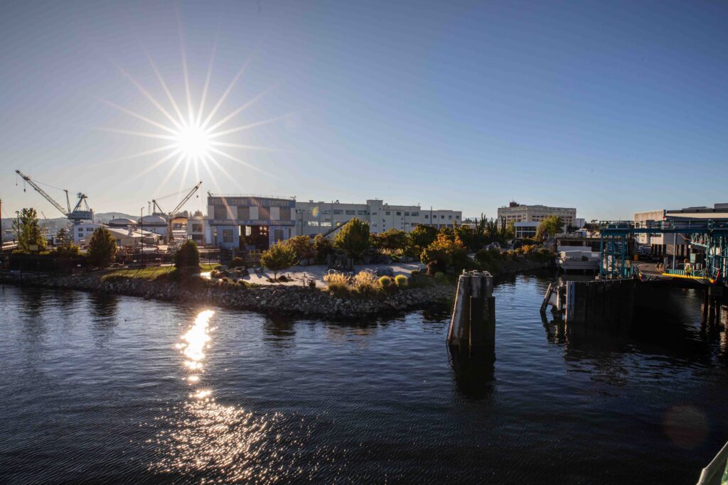 View from a ferry approaching Bremerton, with sunlight reflecting across Sinclair Inlet toward Puget Sound Naval Shipyard, waterfront buildings, cranes, and the ferry terminal.