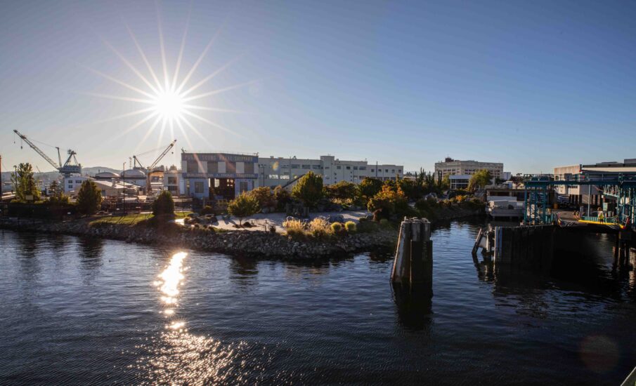 View from a ferry approaching Bremerton, with sunlight reflecting across Sinclair Inlet toward Puget Sound Naval Shipyard, waterfront buildings, cranes, and the ferry terminal.